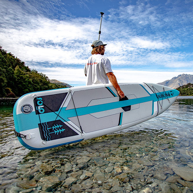 "Pau Hana Surf Supply 10'10" Solo SUP Backcountry inflatable SUP board on alpine lake with mountain backdrop, featuring signature textured deck pad"