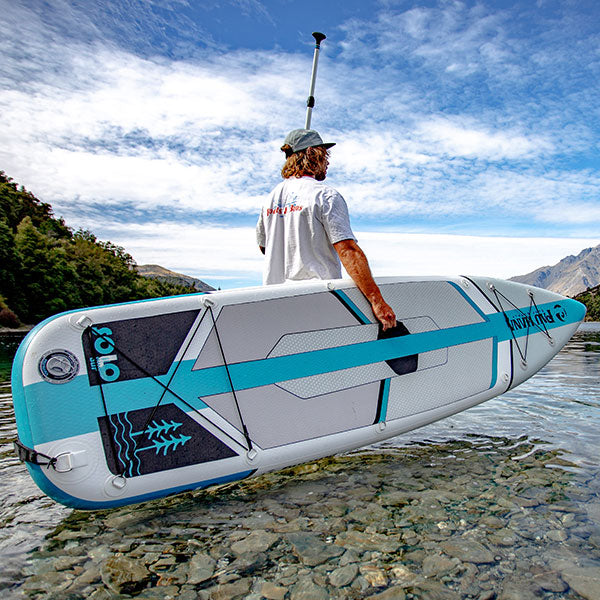 "Pau Hana Surf Supply 10'10" Solo SUP Backcountry inflatable SUP board on alpine lake with mountain backdrop, featuring signature textured deck pad"