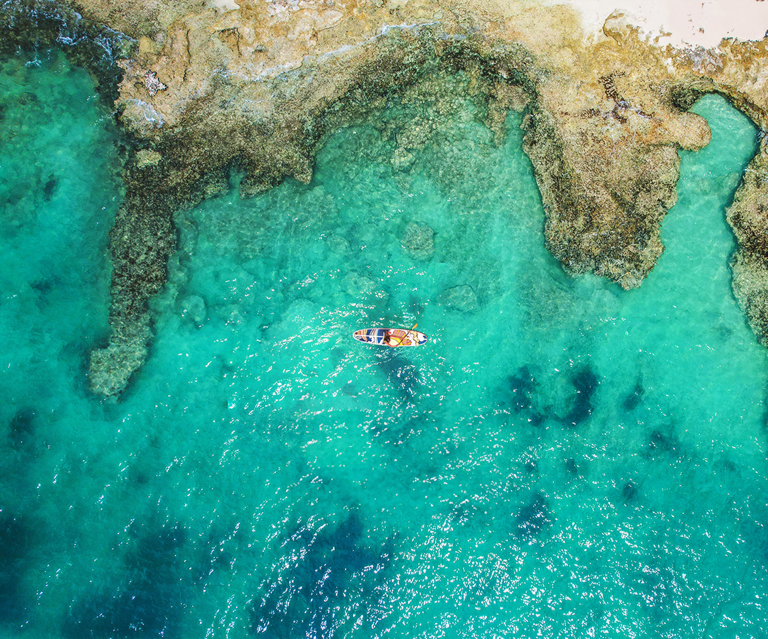 An aerial image of Pau Hana Stowaway Lite inflatable paddleboard artistic design Hippy Wave on the ocean next to an outcrop of rocks