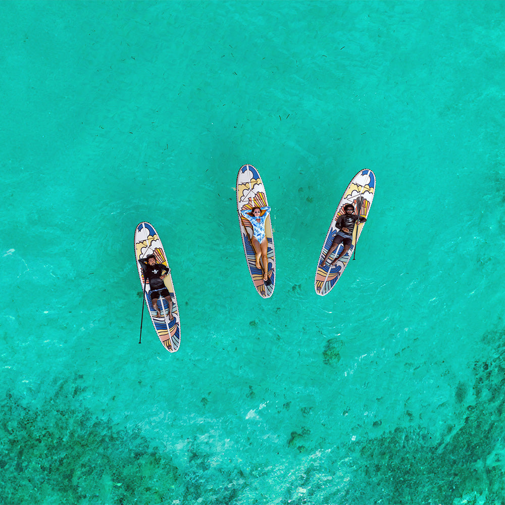 "Aerial drone shot of three paddleboarders on Pau Hana surf Supply Stowaway Lite Hippy Wave design boards, floating over crystal-clear turquoise reef waters"