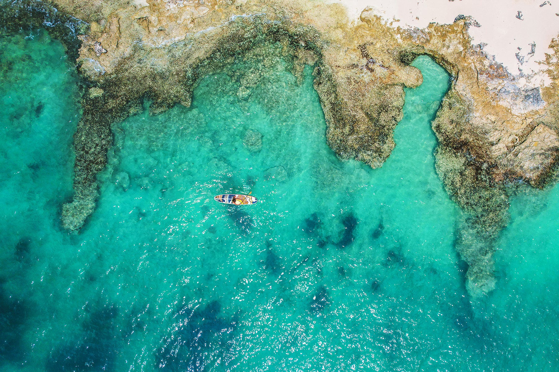 An aerial image of a paddleboard on the ocean next to the shoreline of rocky outcrops