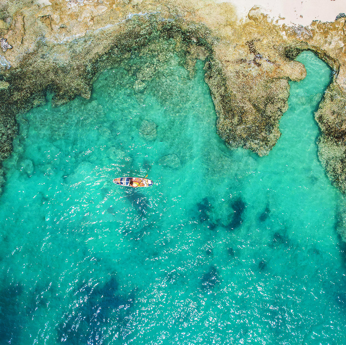 "Aerial drone shot of paddleboarder on Pau Hana Surf Supply Stowaway Lite Hippy Wave art inspired inflatable board, floating over crystal-clear turquoise lagoon waters"