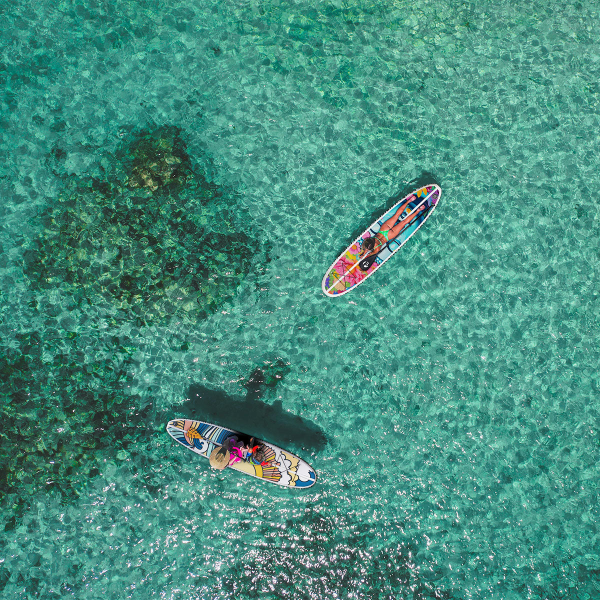 "Aerial drone shot of paddleboarders on Pau Hana Surf Supply Stowaway Lite Hippy Wave and Power of Flower design inflatable boards, floating over crystal-clear turquoise reef waters"