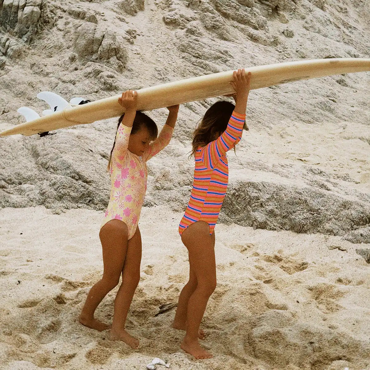 Two children carrying a surfboard on a sandy beach with rocky cliffs in the background.