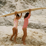 Two children carrying a surfboard on a sandy beach with rocky cliffs in the background.