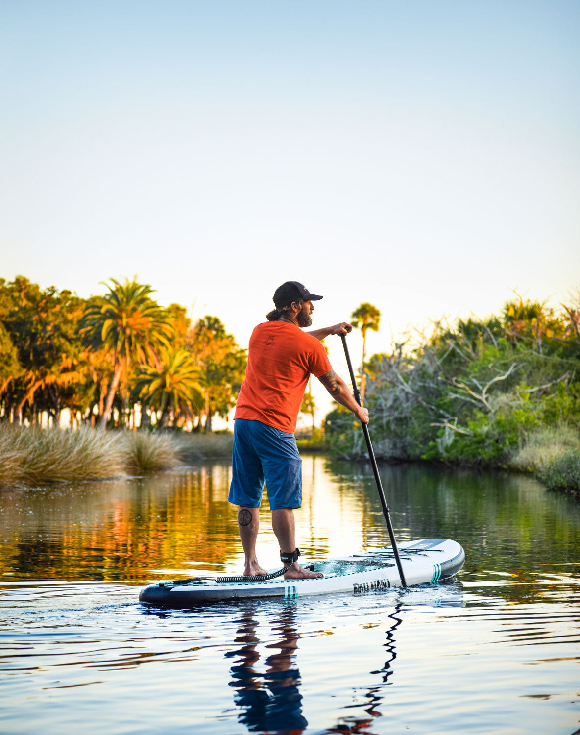 "Paddleboarder on Pau Hana Surf Supply Big EZ Air gliding through calm coastal waters at sunset, surrounded by tropical palms - showcasing all-around touring performance"