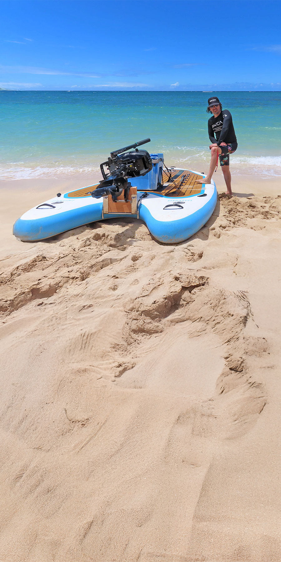 Person standing next to a blue and white inflatable giant paddleboard on a sandy beach with clear blue water and sky.