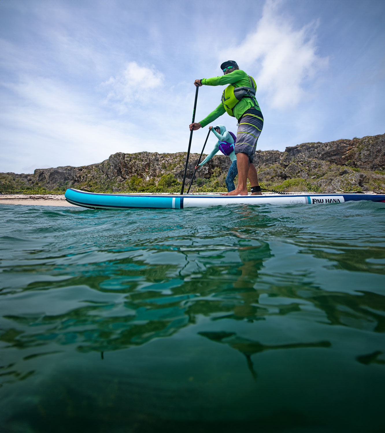 "Paddler in neon green rashguard and PFD navigates Pau Hana Surf 
Supply Calypso touring inflatable SUP along rocky coastline, shot from water level showing clear turquoise ocean depths"