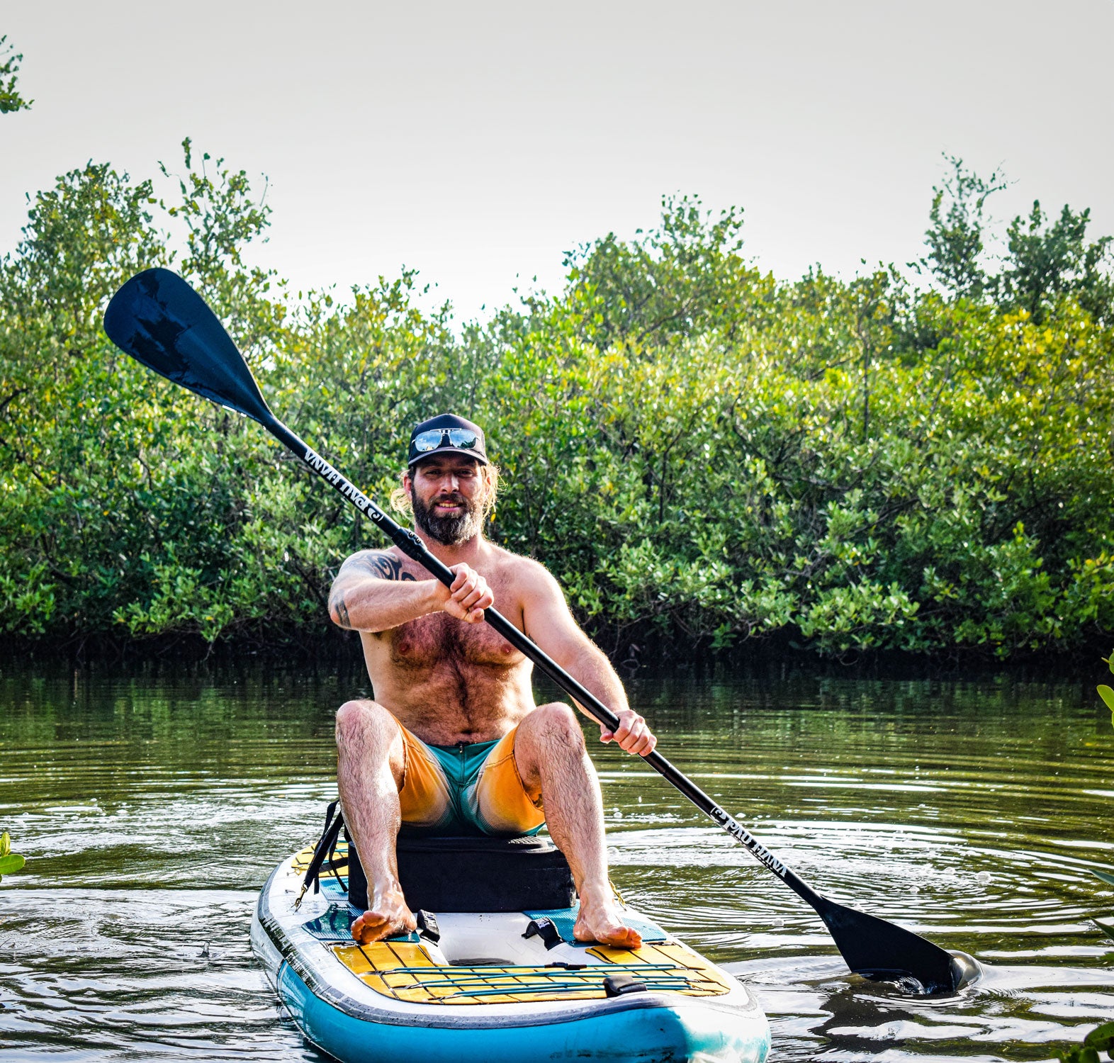Man paddleboarding on a calm body of water with greenery in the background
