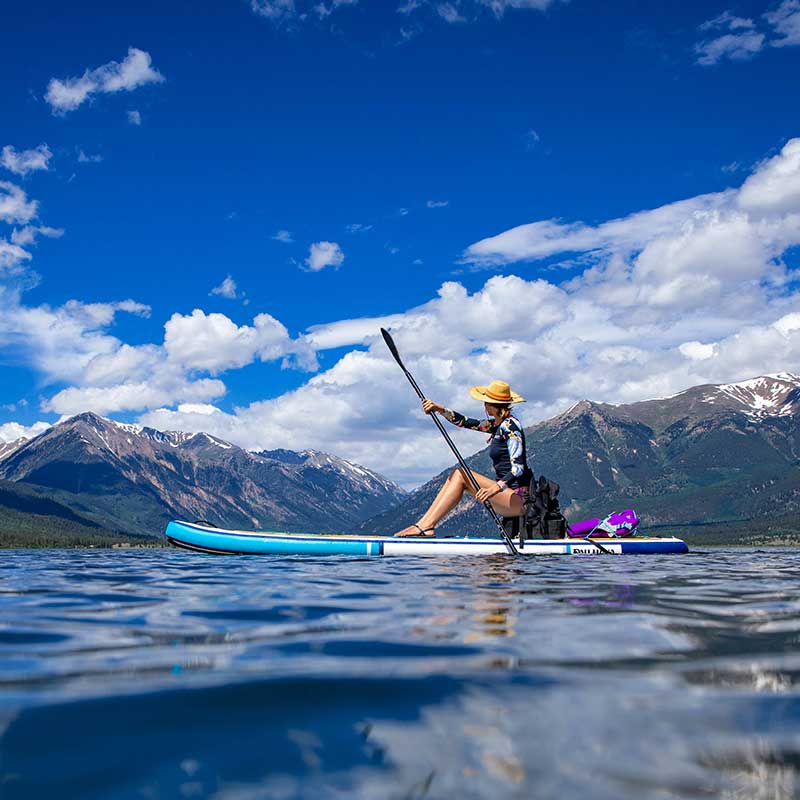 "Pau Hana Surf Supply Calypso inflatable SUP with kayak conversion kit on alpine lake, paddler using convertible paddle beneath mountain peaks"