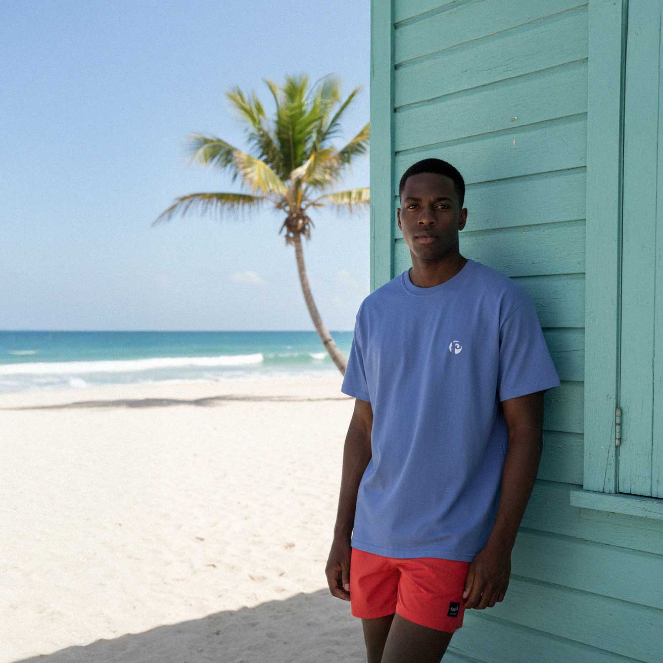 Man in a blue shirt and red shorts standing by a teal building with a palm tree and ocean in the background.