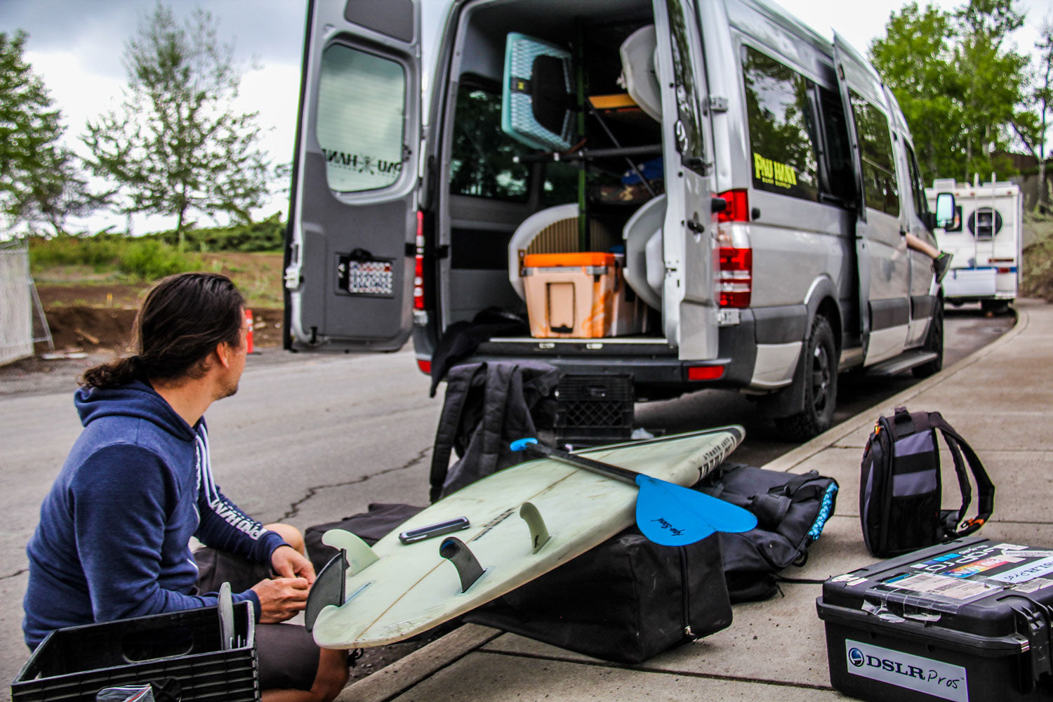 "Pau Hana Surf Supply team member prepping Carve surf SUP by adventure van, equipped with blue paddle and installing fins"