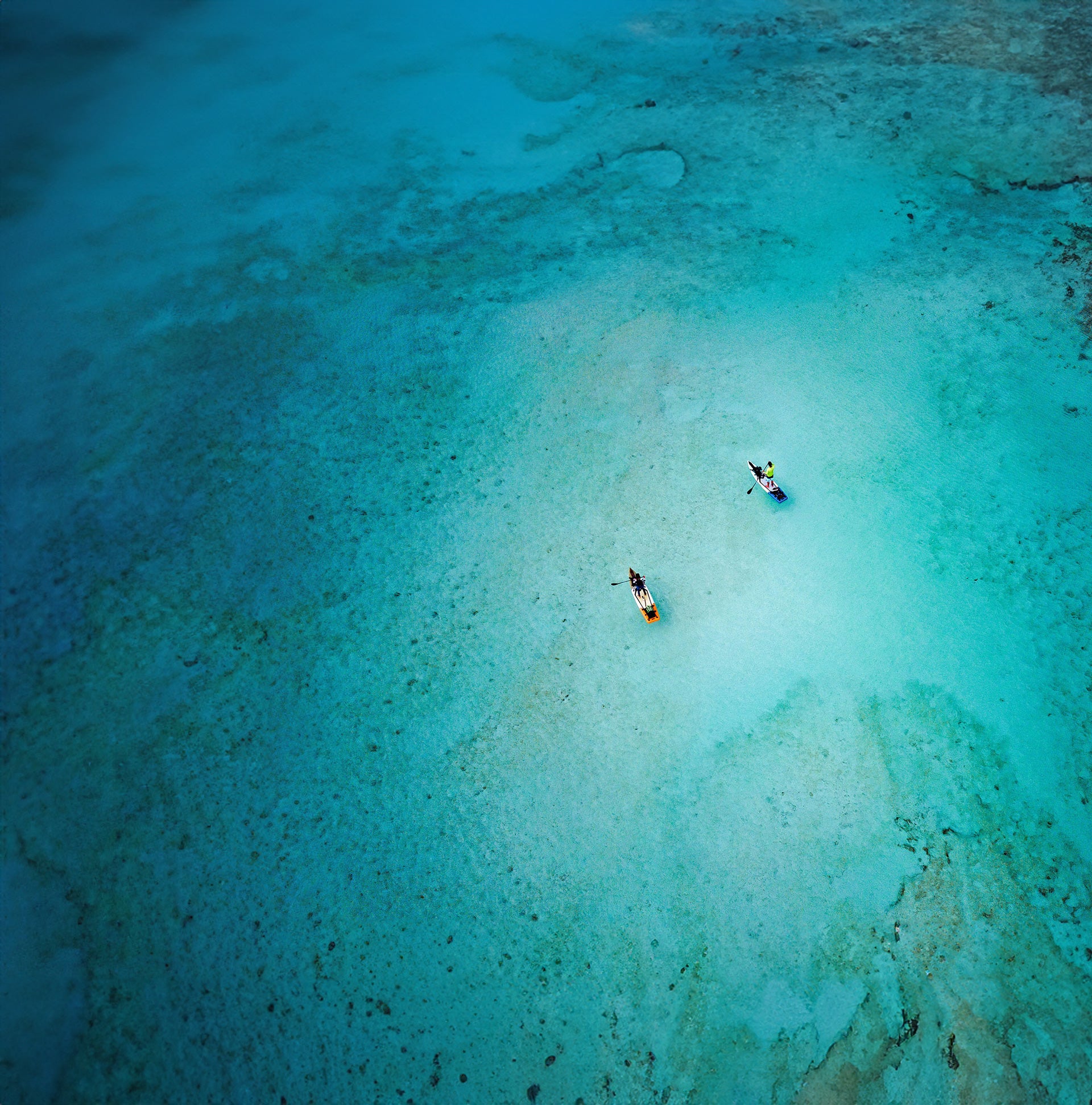 "Aerial drone shot of paddleboarders on Pau Hana Surf Supply Endurance Air inflatable touring boards, loaded with camping gear, floating over crystal-clear turquoise reef waters"