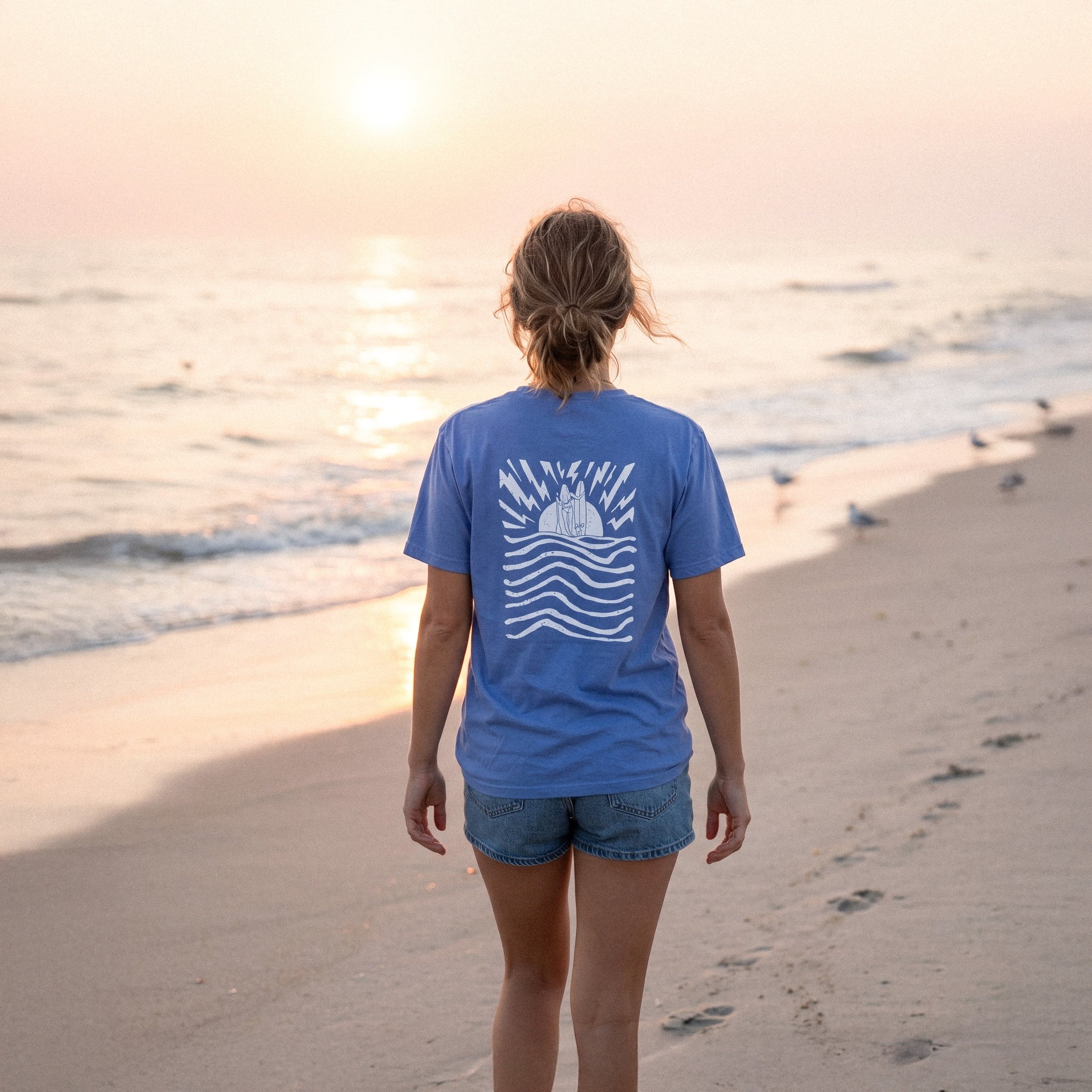 Person wearing a blue t-shirt with a graphic design on the beach at sunset.