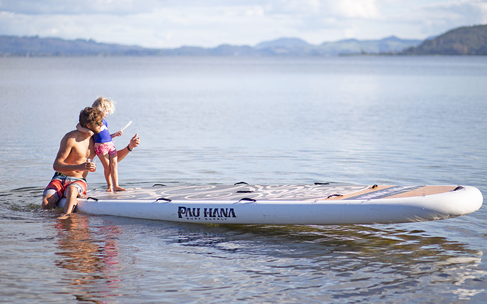 "A family enjoys time on the Pau Hana Surf Supply Oahu Nui giant family inflatable SUP in serene New Zealand lake surrounded by mountains"