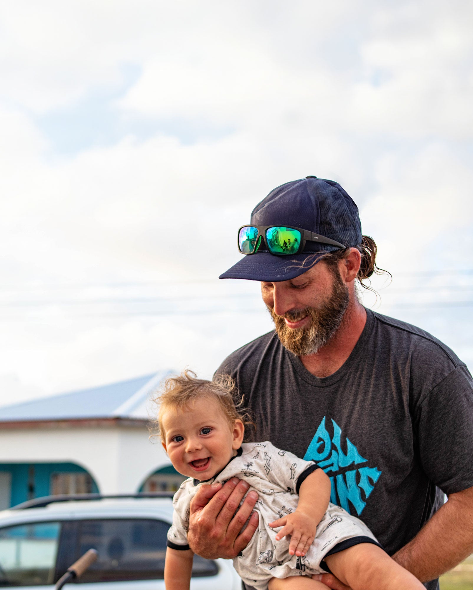 "Pau Hana team member sharing joyful moment with smiling toddler, wearing company surf apparel and mirrored sunglasses outdoors"