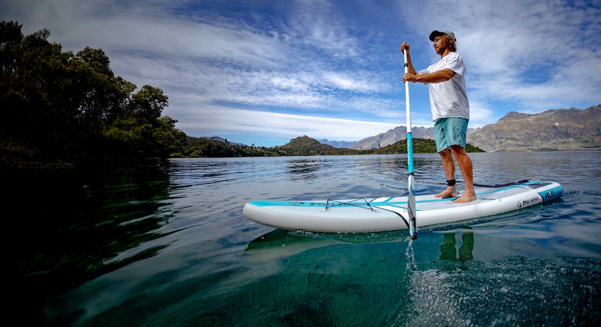A man stands on a white and teal stand-up paddleboard on calm blue water, holding a paddle. Trees and mountains are visible in the background under a partly cloudy sky.