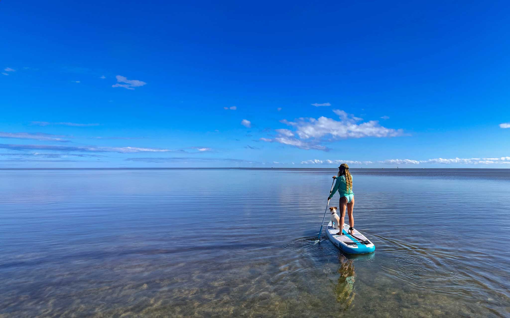 A woman stands on a paddleboard on calm blue water under a bright blue sky, viewed from behind as she paddles in an open lake or sea.