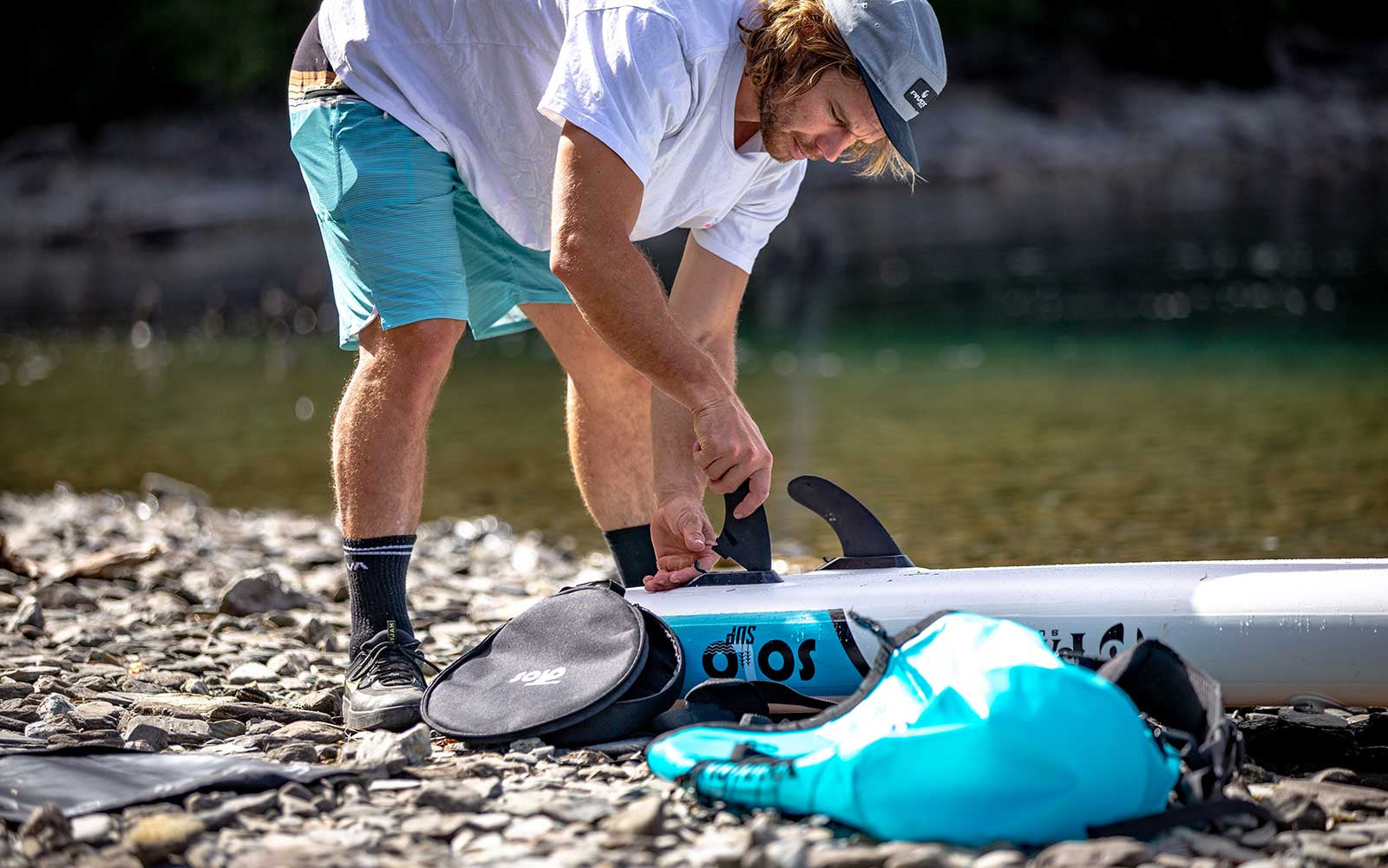 Paddler installing snap-lock fins on Pau Hana Surf Supply SOLO SUP board at rocky shoreline, showcasing quick-connect fin system setup process