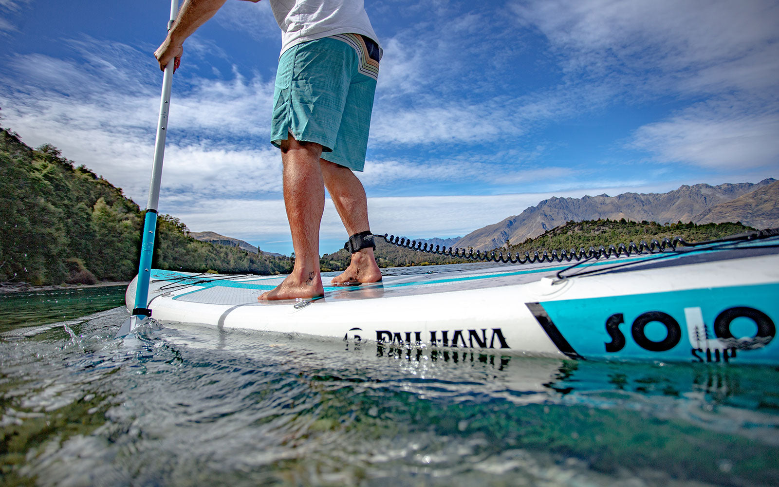 Pau Hana Surf Supply SOLO SUP Backcountry on alpine lake with mountain backdrop, showing coiled leash and EZ-Adjust paddle during backcountry adventure